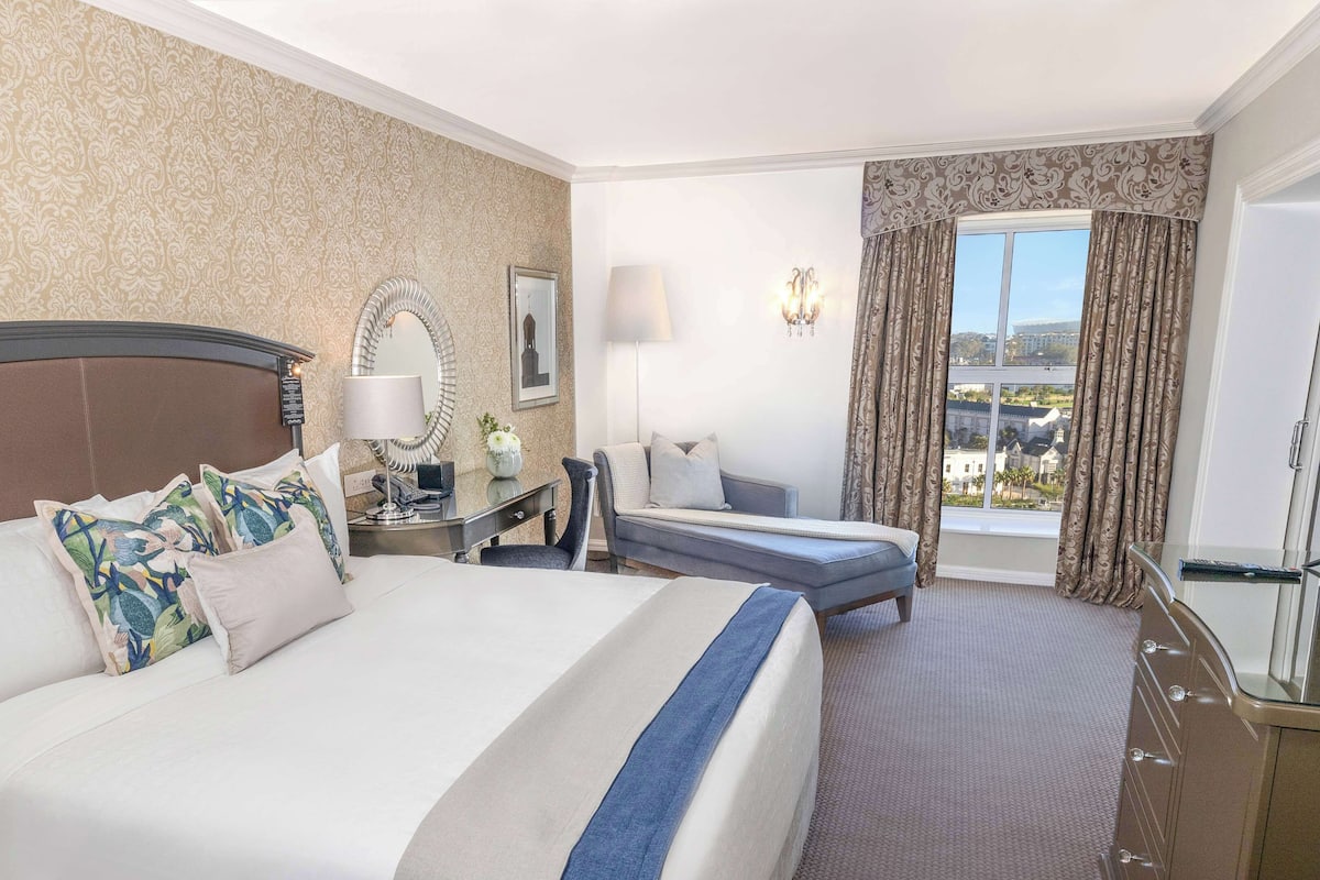 A hotel room with a large bed, a desk with a lamp, a chair, a sofa, and a view of the city through the window.