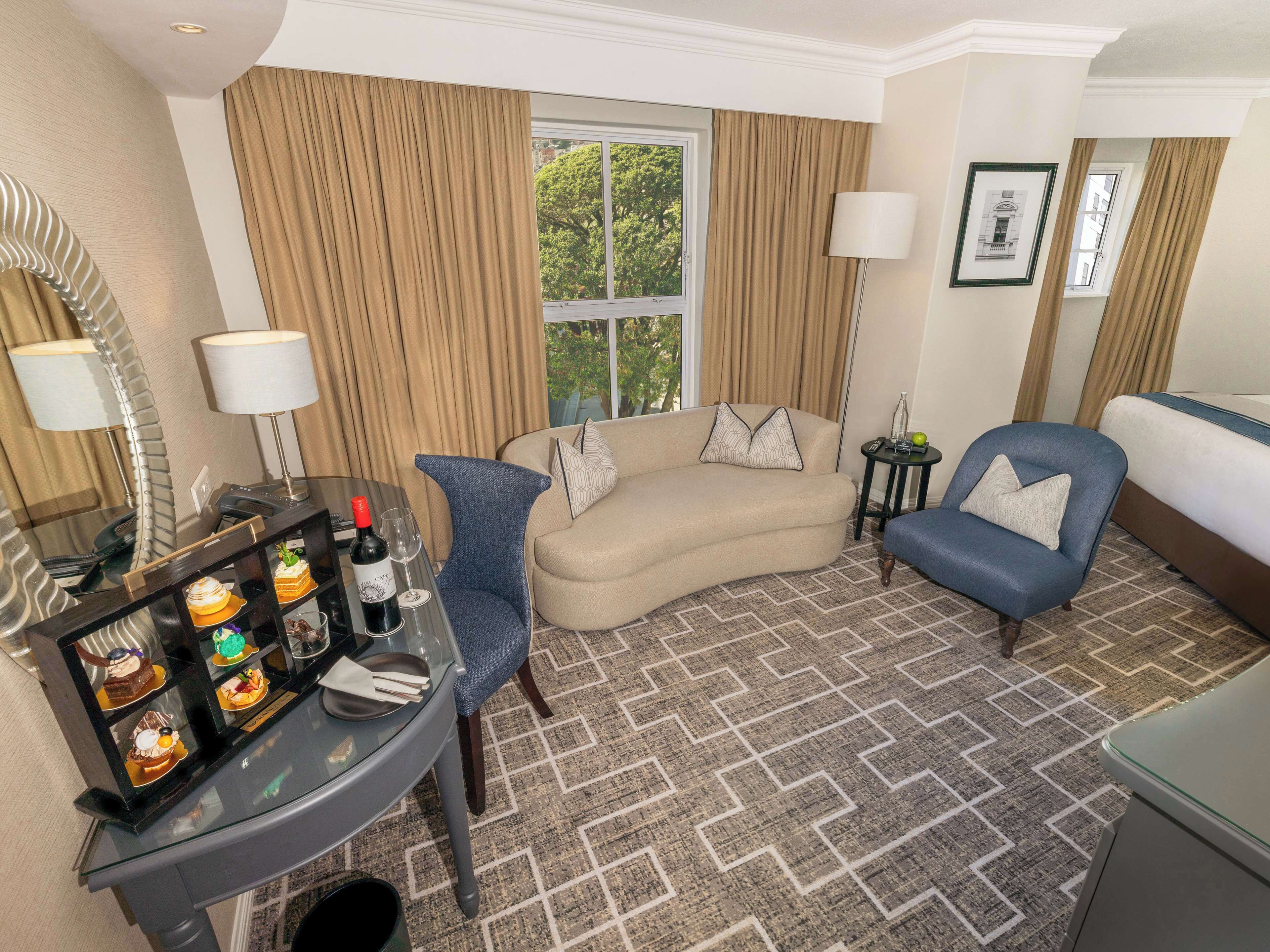 A hotel room with a curved sofa, a blue armchair, a small table with a bottle and glasses, and a decorative glass display case.