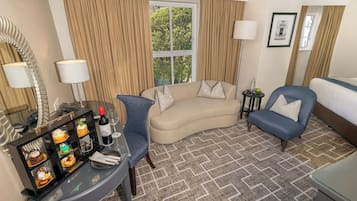 Habitación de hotel con sofá curvo, un sillón azul, una mesita con una botella y vasos, y una vitrina decorativa de vidrio.