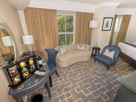 A hotel room with a curved sofa, a blue armchair, a small table with a bottle and glasses, and a decorative glass display case.