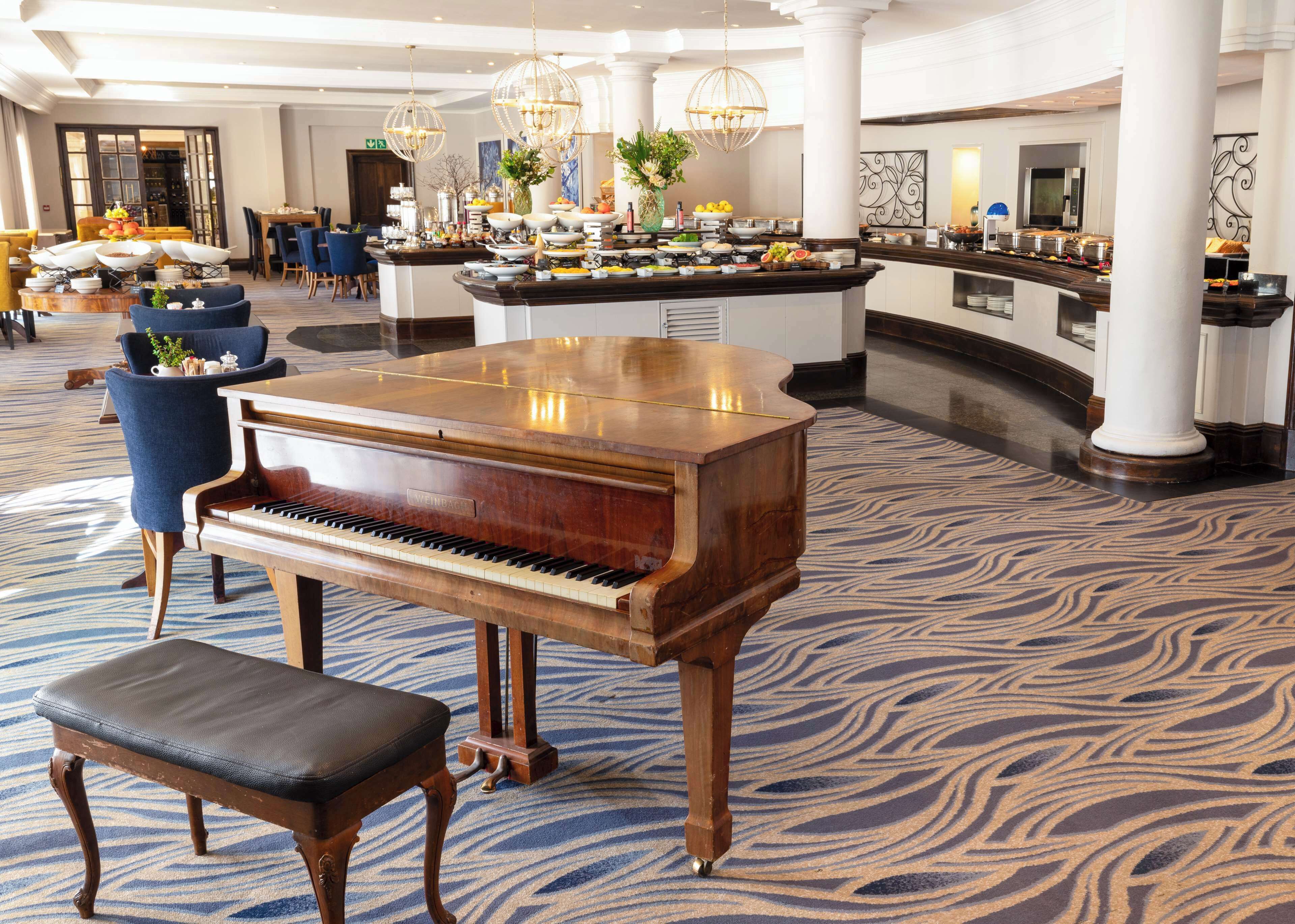A grand piano in a spacious lobby with a patterned carpet, a buffet area with various food items, and a seating area with chairs and a table.