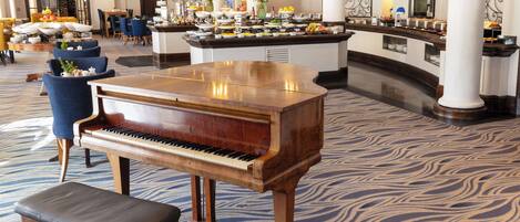 A grand piano in a spacious lobby with a patterned carpet, a buffet area with various food items, and a seating area with chairs and a table.