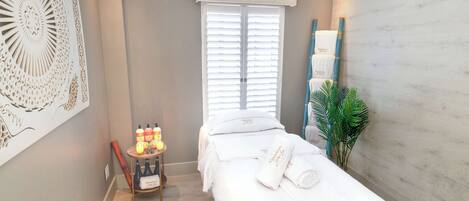 Un cuarto de spa con una camilla de masaje, una mesita con velas y botellas, y una decoración grande en la pared.