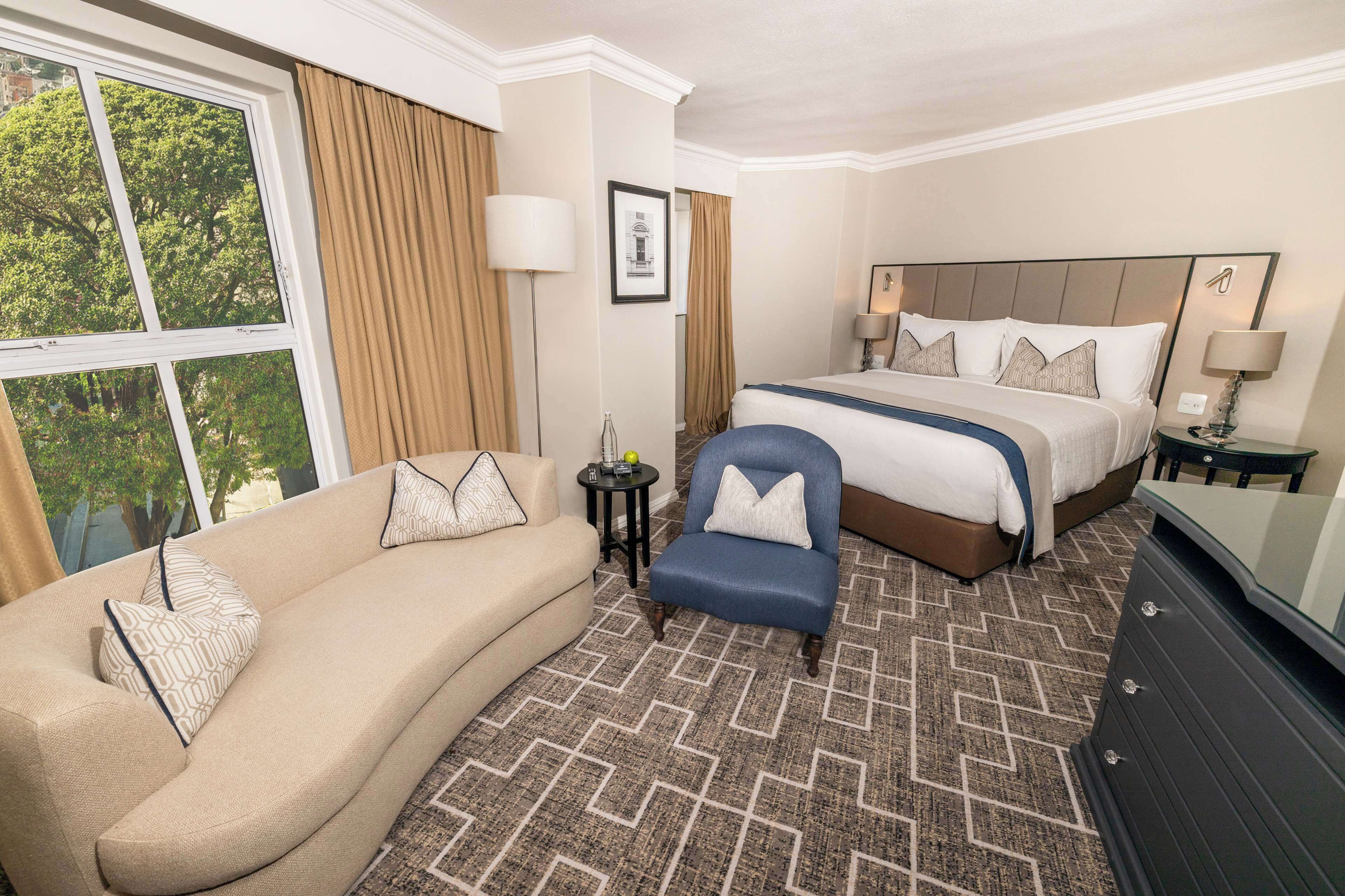 A hotel room with a large bed, a sofa, a chair, a dresser, and a window with curtains.