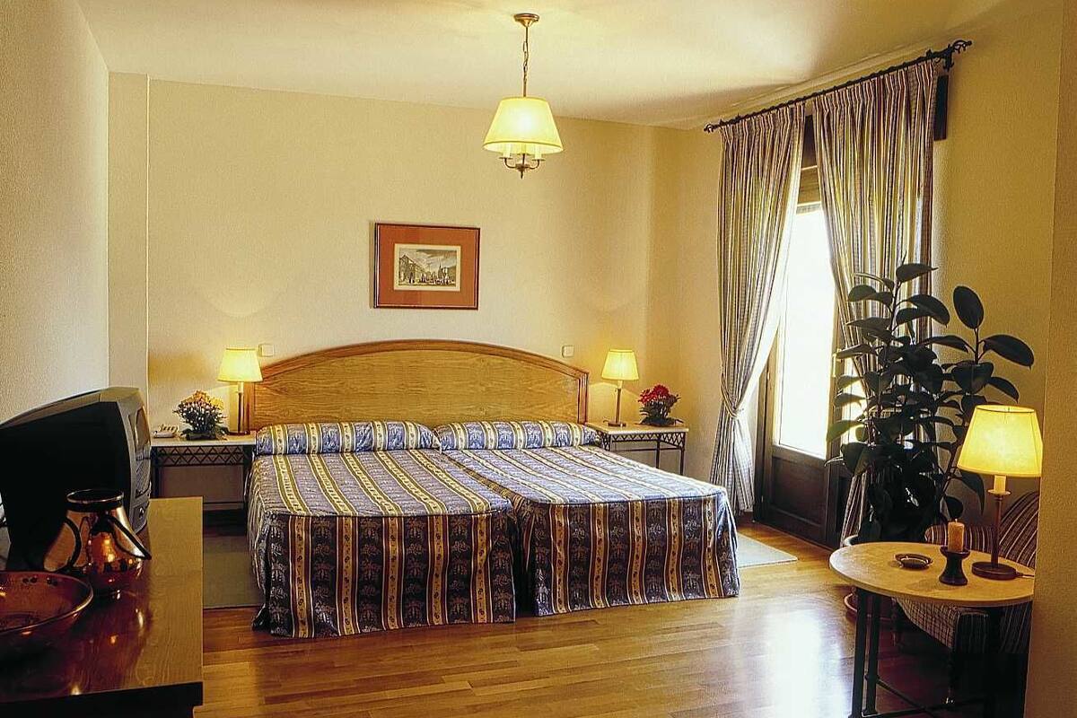 Ein Hotelzimmer mit einem hölzernen Bett, zwei Nachttischen mit Lampen, einem Schreibtisch mit Stuhl, einer Zimmerpflanze und einem Fenster mit Vorhängen.