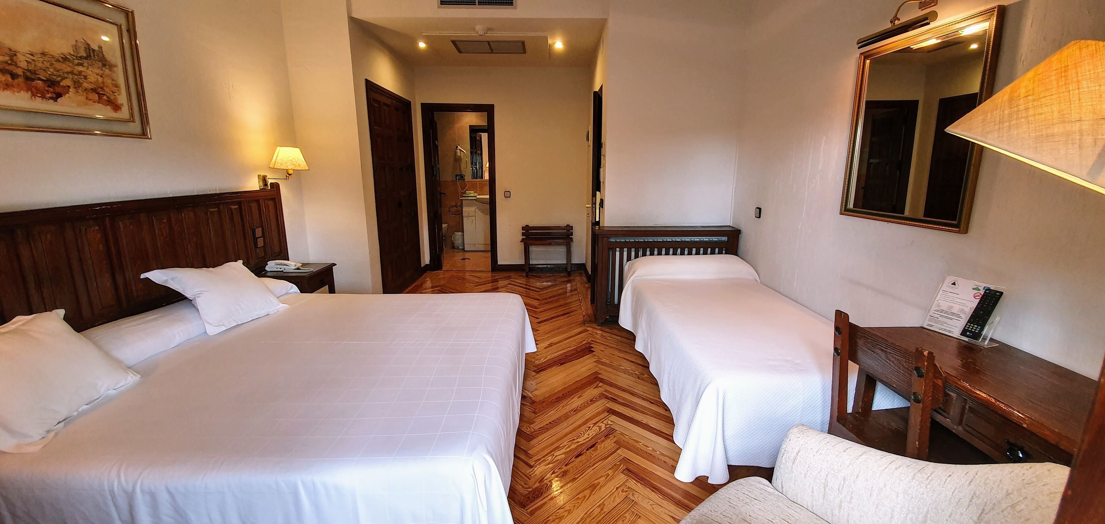 A hotel room with two beds, wooden flooring, a desk, a chair, a lamp, a mirror, and a painting on the wall.