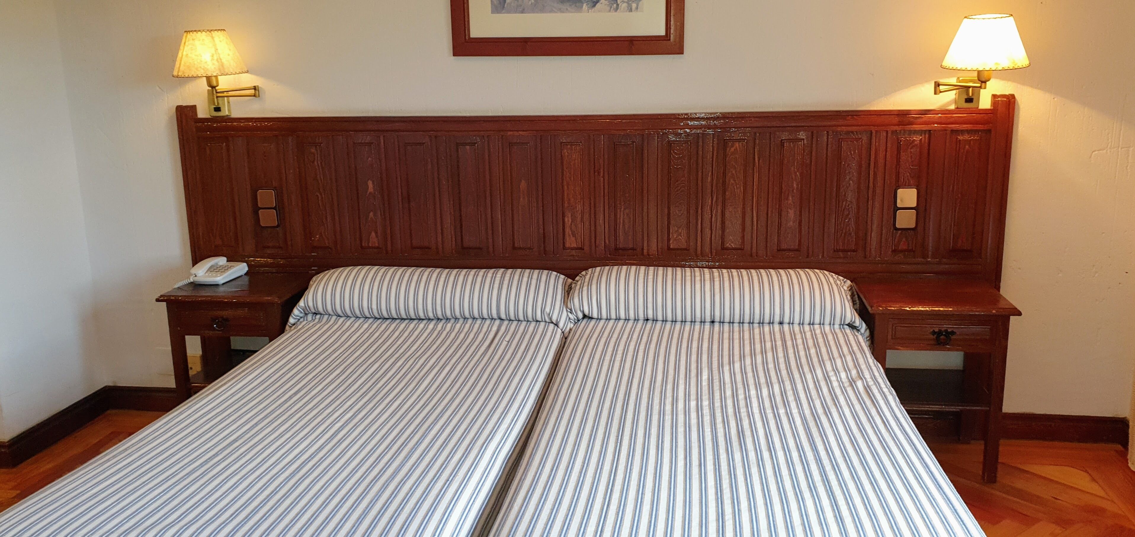 Cama doble con ropa de cama a rayas, cabecero de madera, lámparas de noche, un teléfono y un cuadro en la pared.