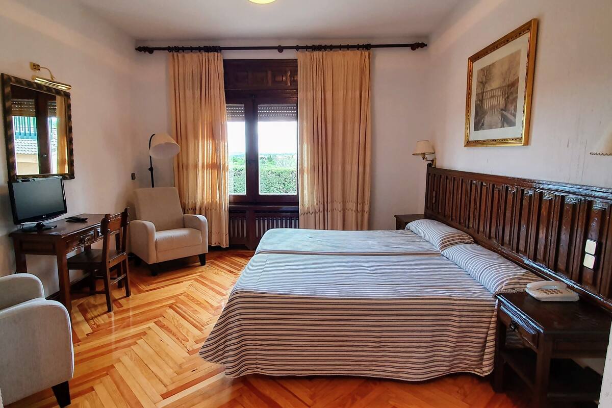 Ein Hotelzimmer mit einem großen Bett, einem Schreibtisch mit Computer, einem Sessel und einem Fenster mit Vorhängen.