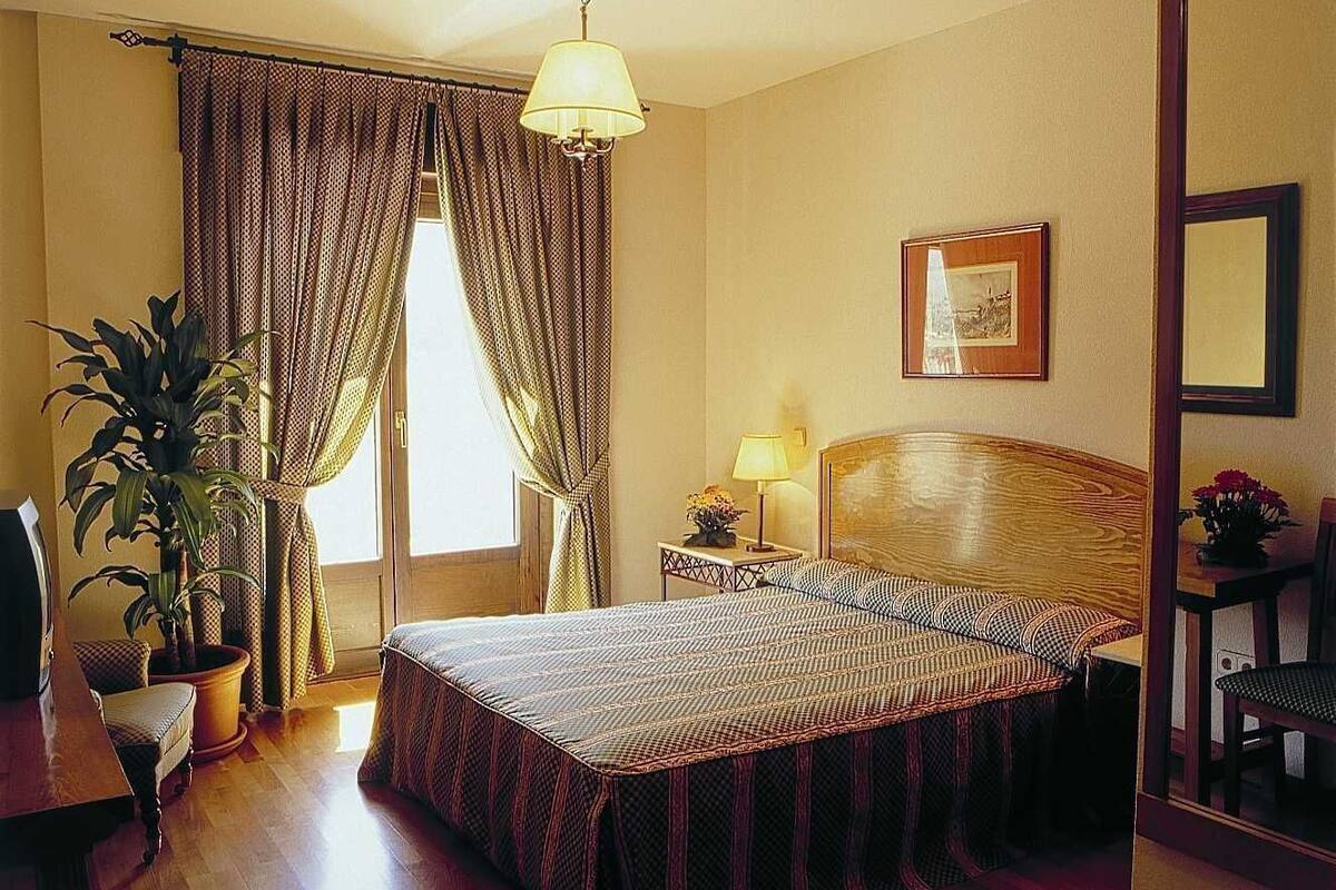 Ein Hotelzimmer mit einem Bett, einem Nachttisch mit Lampe, einem Sessel, einer Zimmerpflanze und einem Fenster mit Vorhängen.