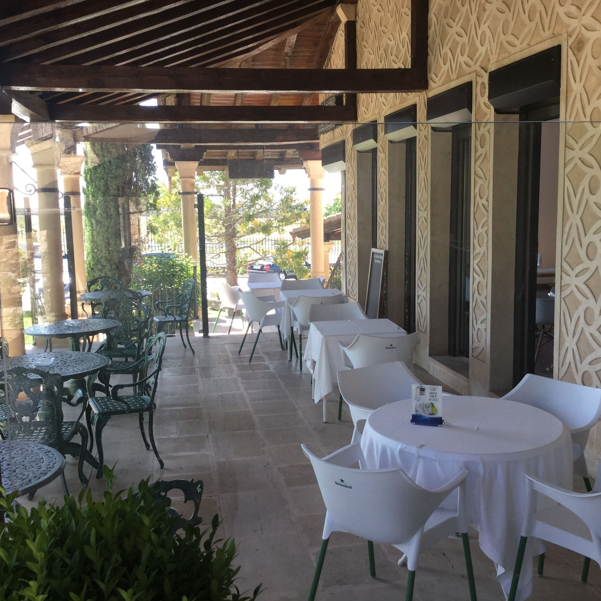 An outdoor patio area with tables and chairs, some covered with white tablecloths, and a decorative wall with intricate patterns.