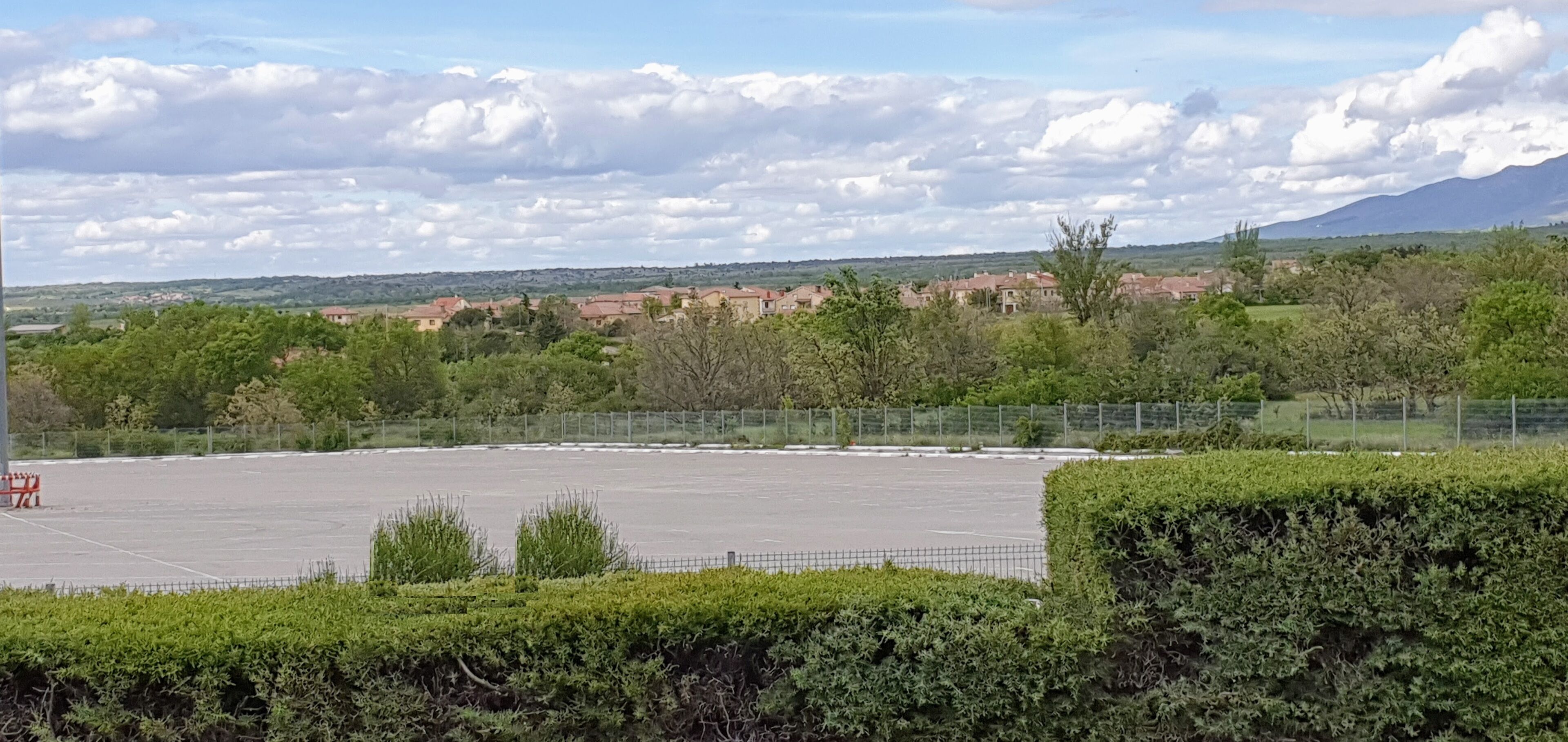 An open parking area with a fence, surrounded by greenery and buildings in the background.