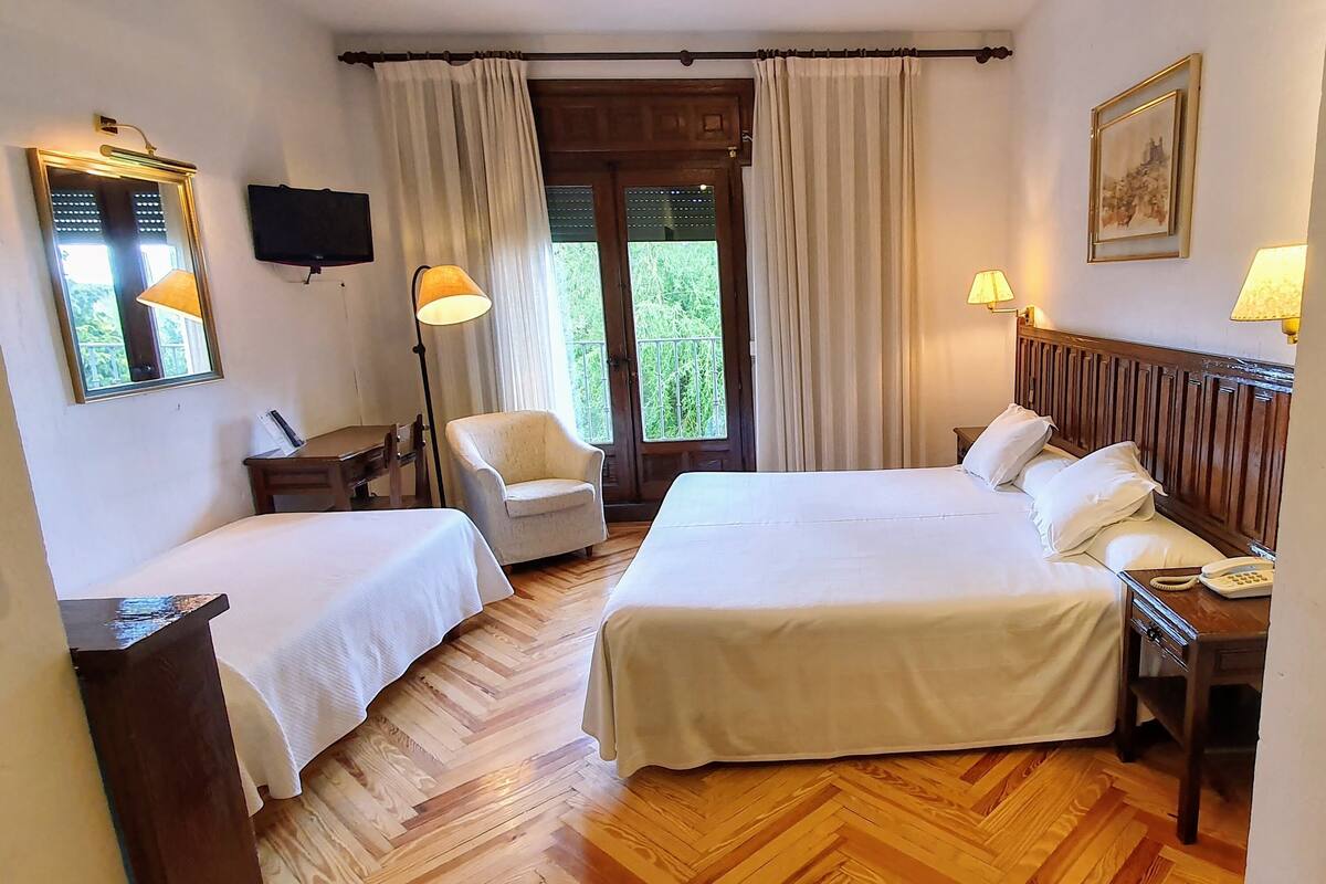 Ein Hotelzimmer mit zwei Einzelbetten, einem Holzboden im Fischgrätmuster, einem Sessel, einem Schreibtisch mit Lampe, einem an der Wand montierten Fernseher und einem Fenster mit Vorhängen.
