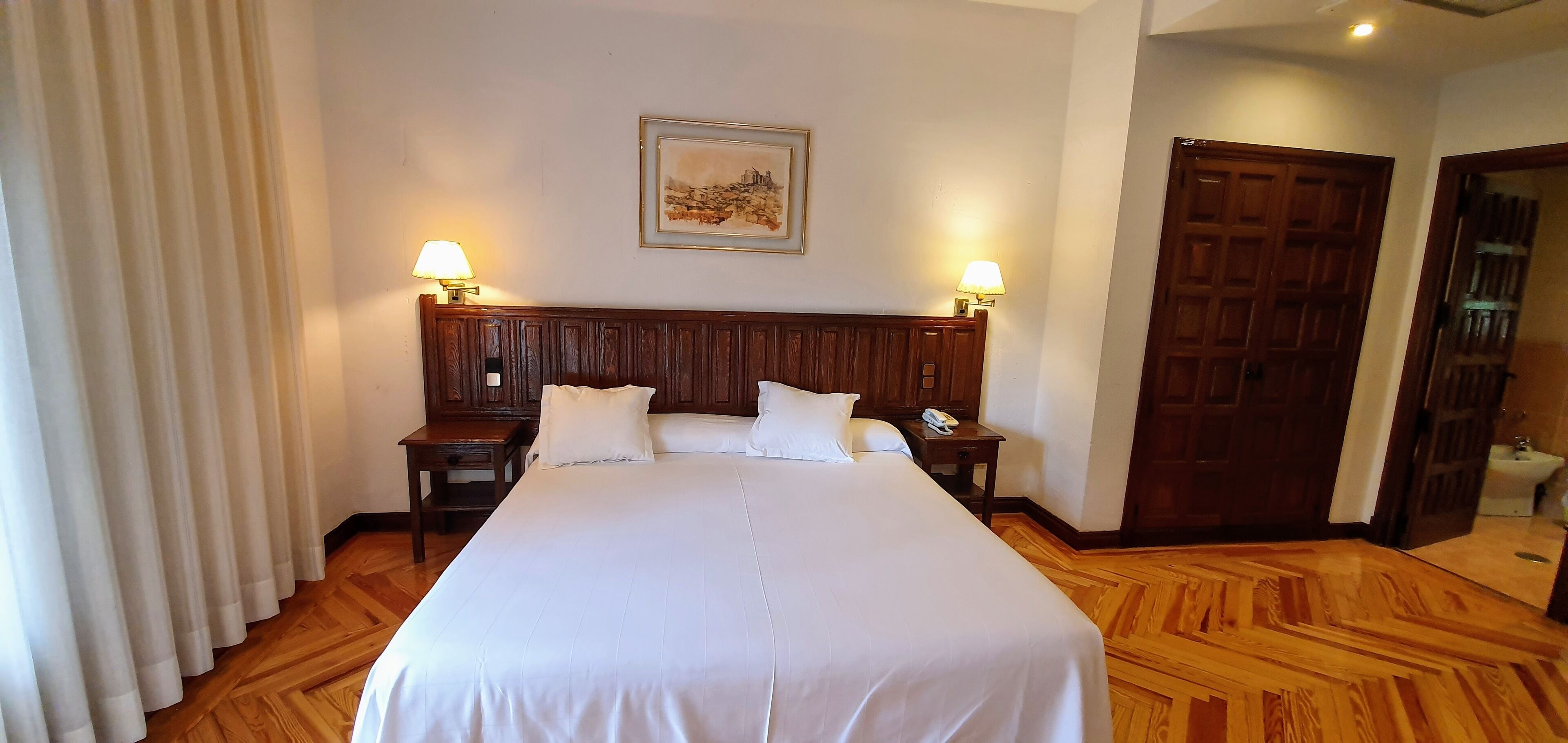 A hotel room with a large bed, two bedside tables with lamps, a wooden headboard, a framed picture on the wall, and a wooden floor.