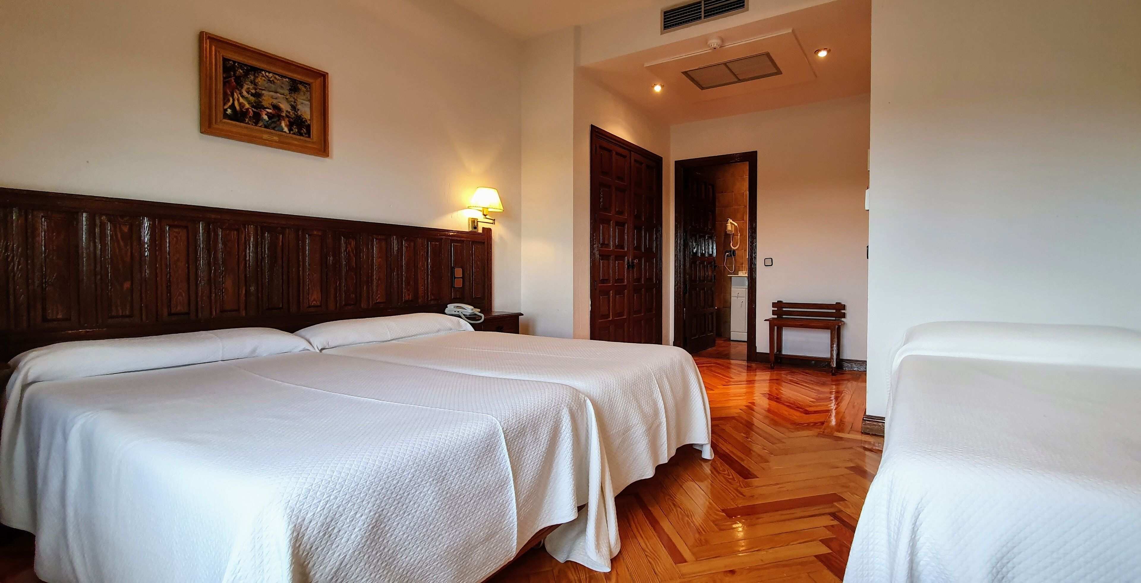 A hotel room with two beds, wooden headboard, a painting on the wall, a bench, and a door leading to a bathroom.