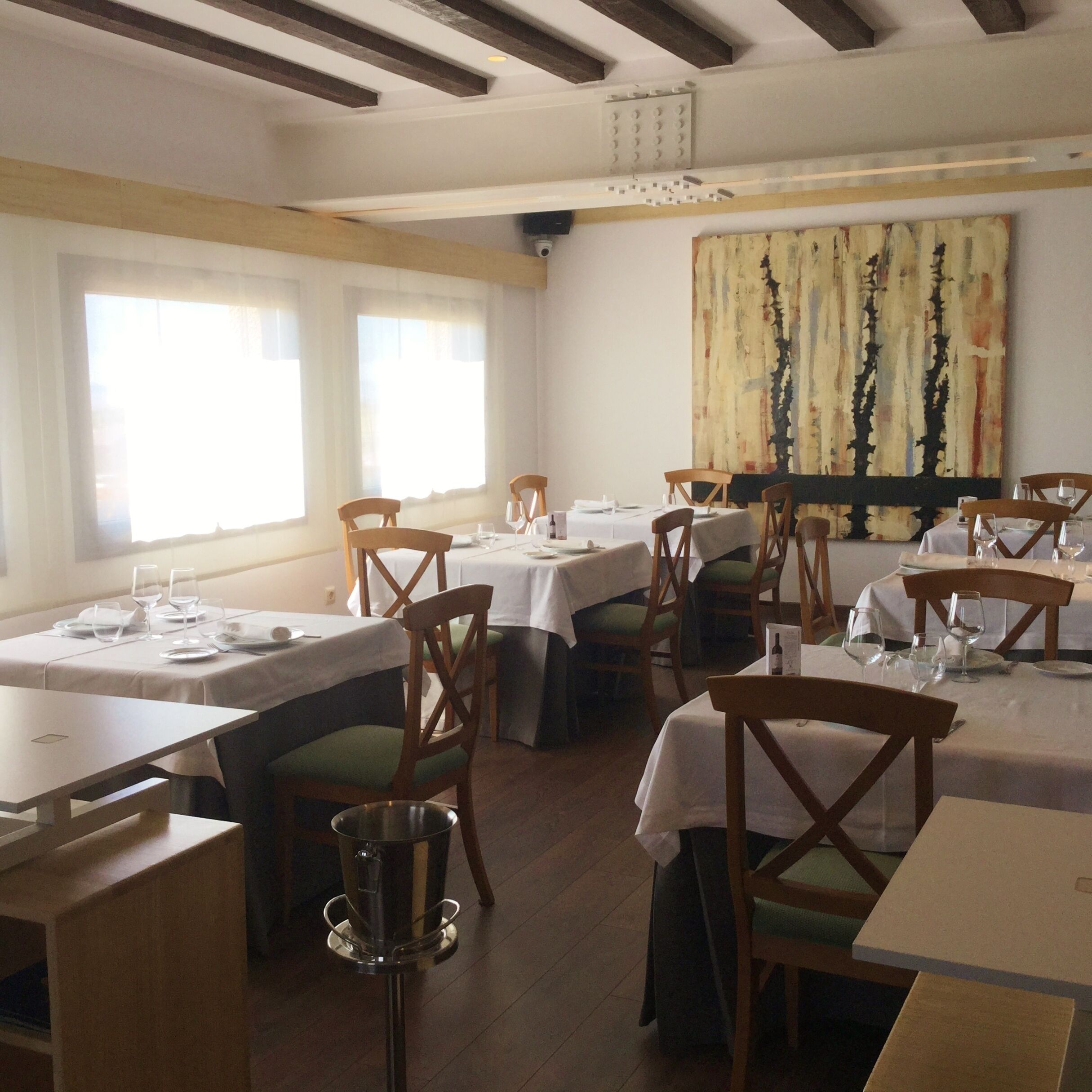 A restaurant with multiple tables and chairs, large windows, and a painting on the wall.