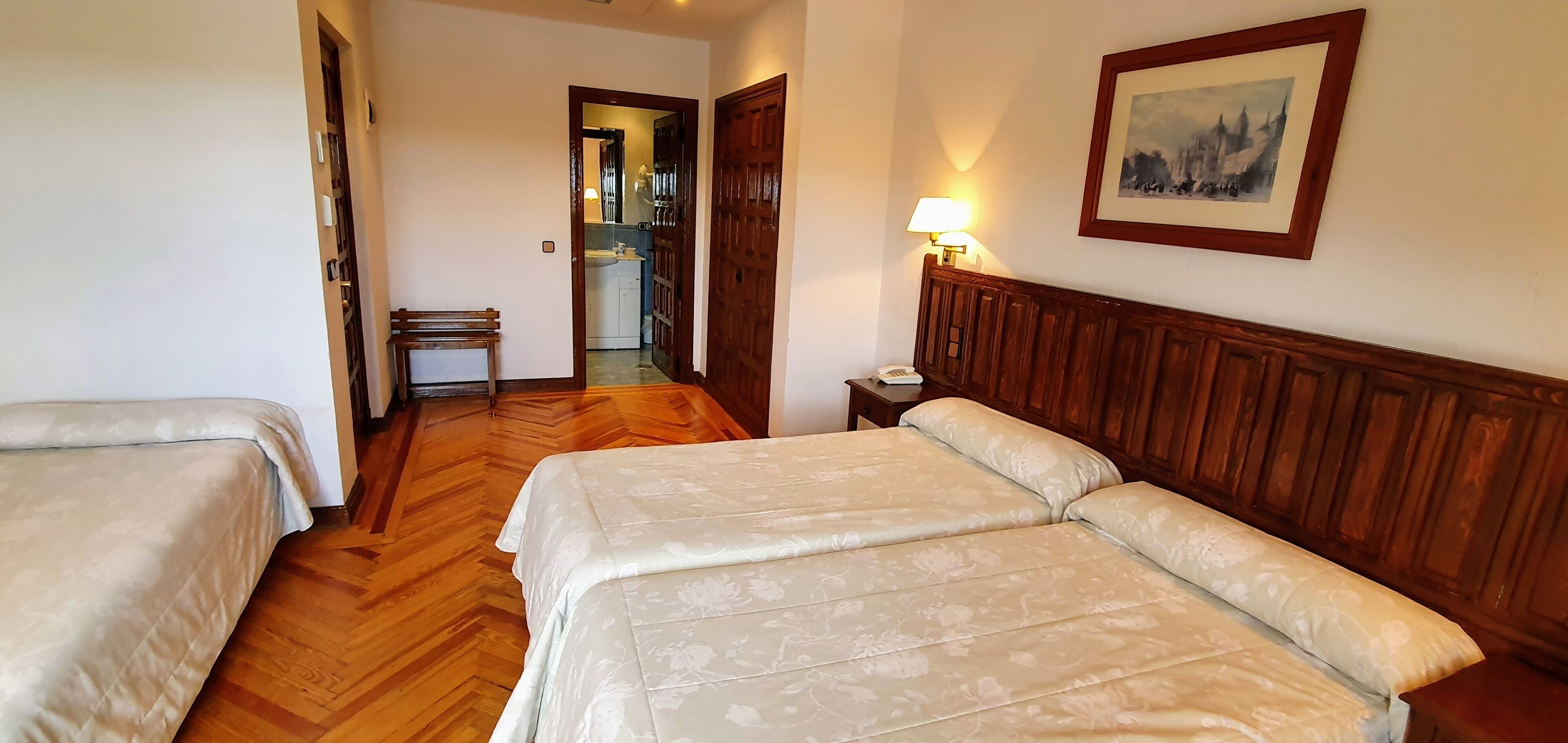 A hotel room with two beds, a wooden headboard, a bedside table with a lamp, and a framed picture on the wall.