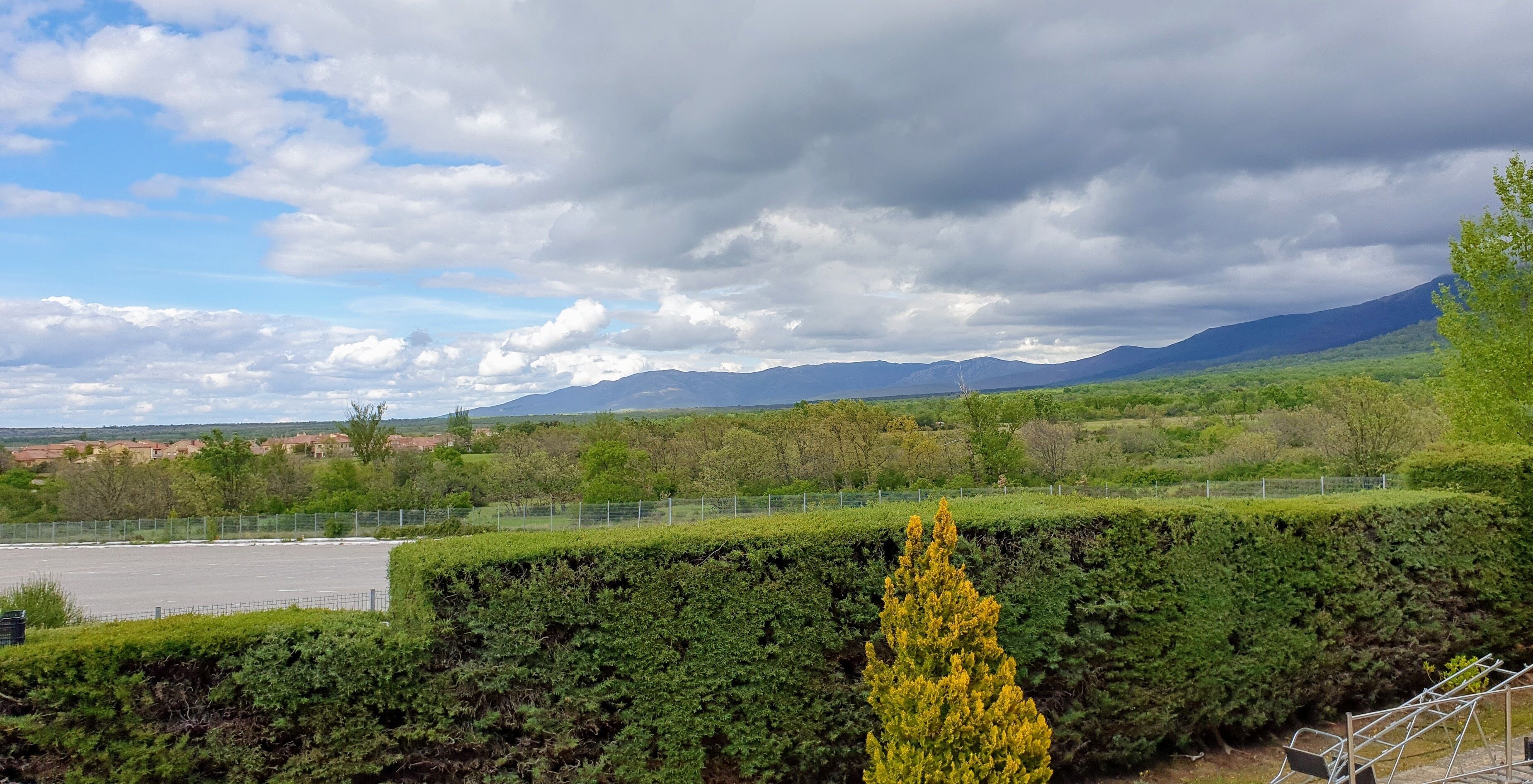 A landscape with a fenced parking area, green hedges, and distant mountains under a partly cloudy sky.