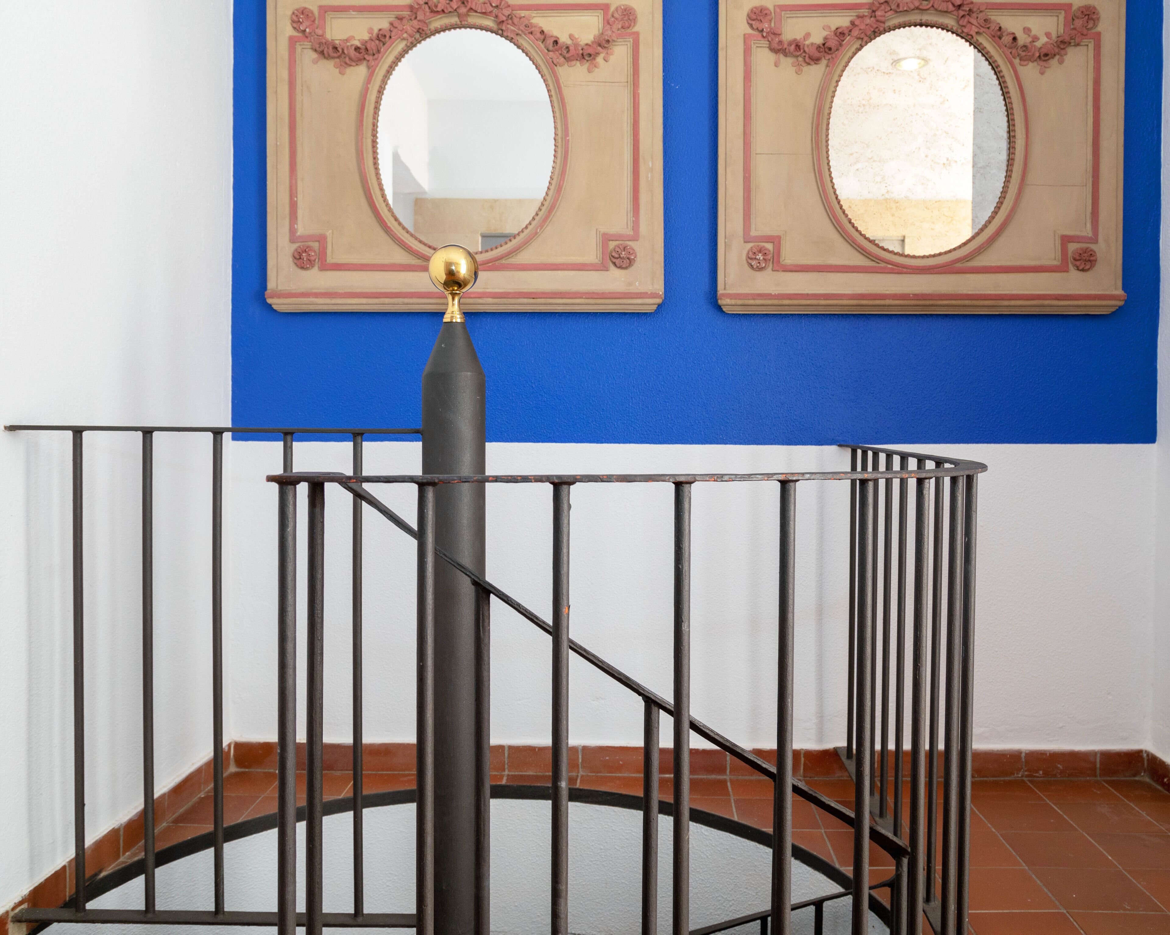 A spiral staircase with a black railing and a golden finial, leading to a room with blue walls and two ornate mirrors.