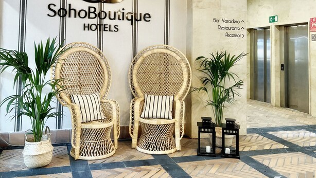 Een hotel lobby met een SohoBoutique Hotels bord, rieten stoelen, potplanten en een betegelde vloer.