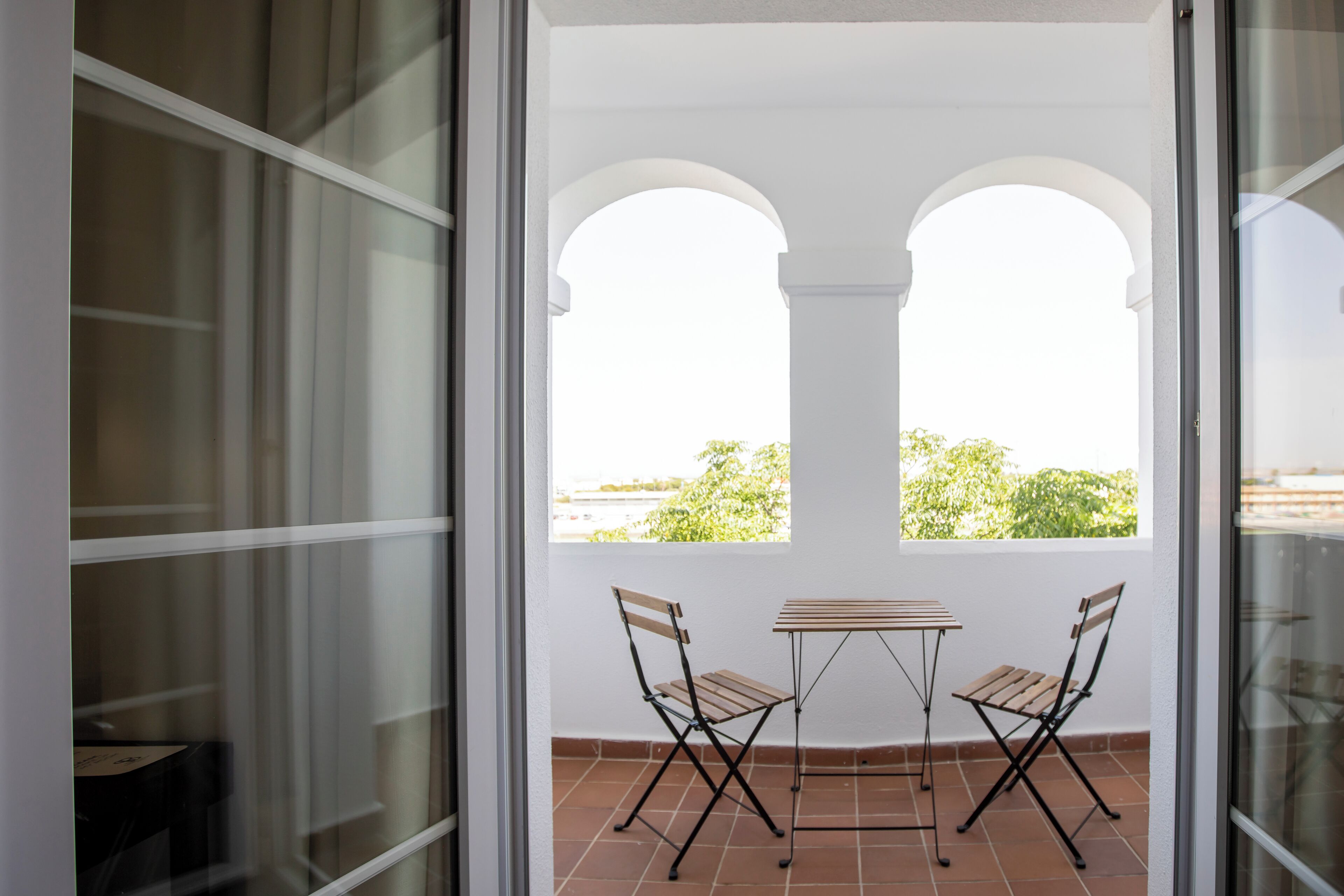 A balcony with two wooden chairs and a small table, overlooking a green landscape through a white archway.