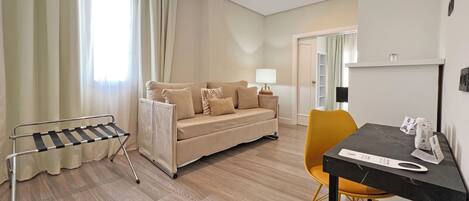Una habitación de hotel moderna con un sofá, una mesa de comedor y una silla. Hay una ventana con cortinas, una lámpara sobre el sofá y un perchero.