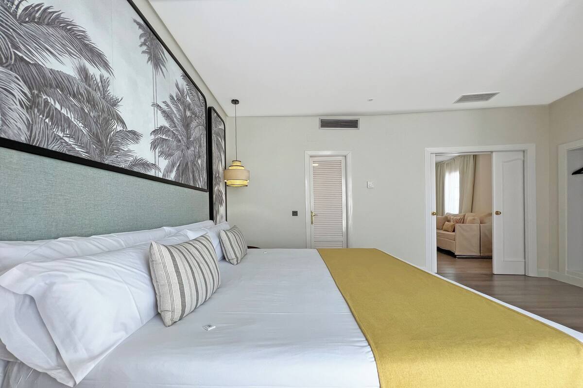 Habitación de hotel con cama, un cuadro de palmeras, una puerta y vistas a otra habitación.