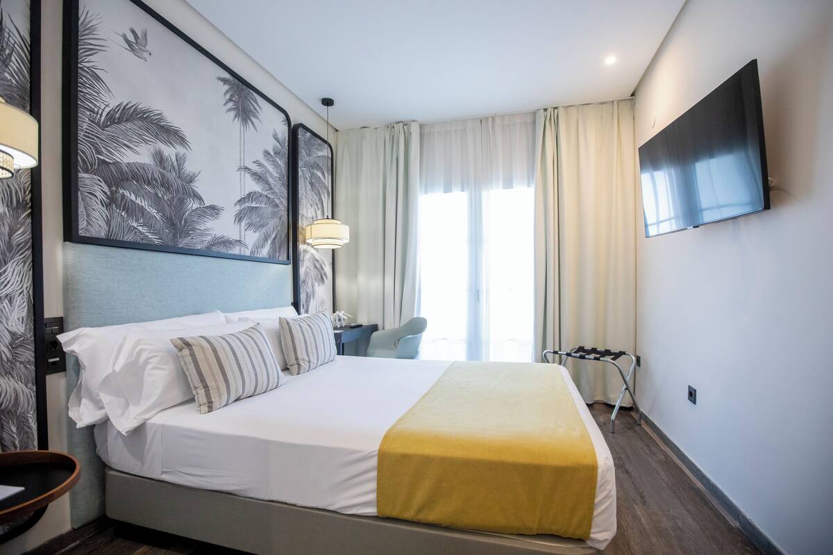 Habitación de hotel con cama, televisor montado en la pared, escritorio con silla y ventana con cortinas.
