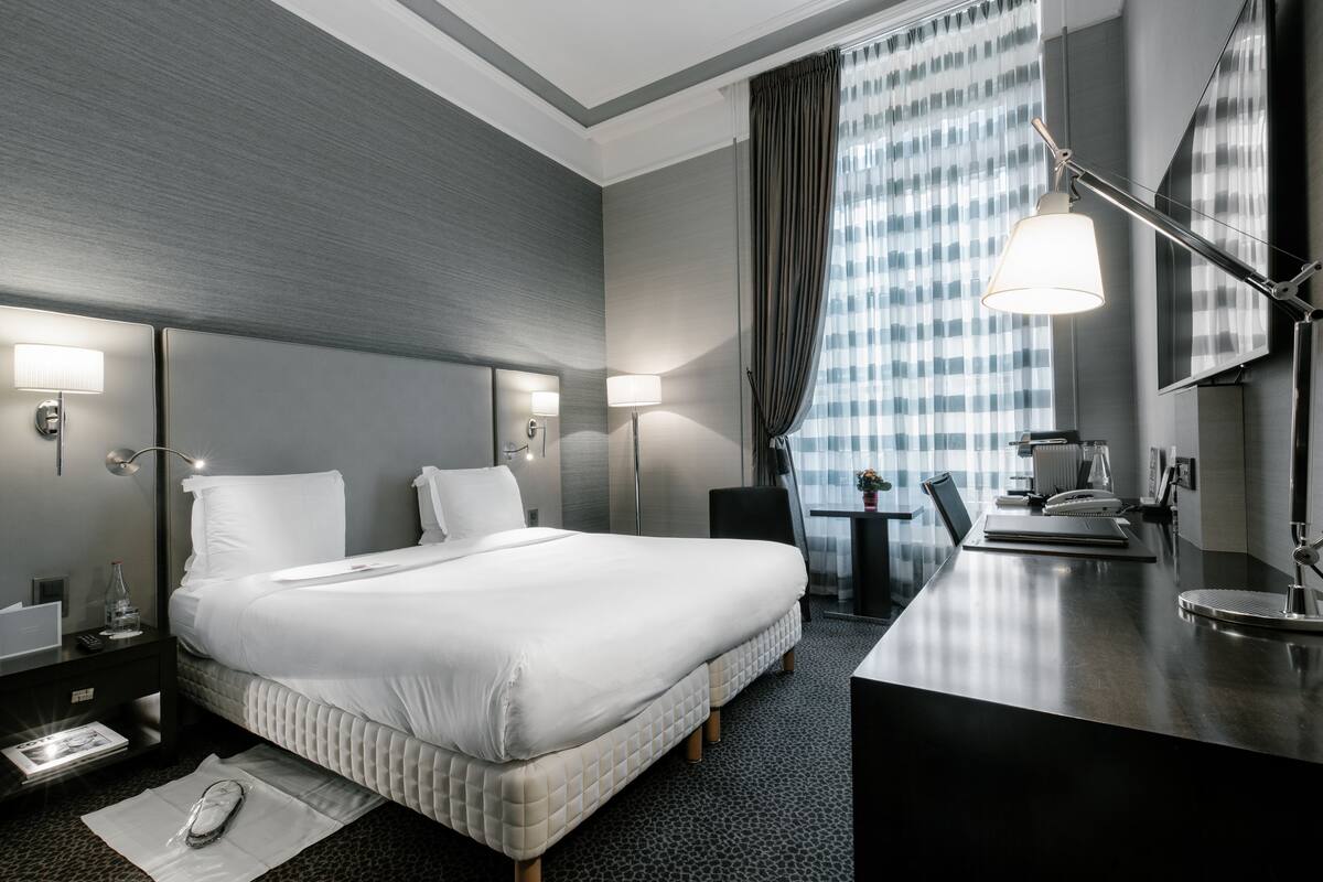 Una habitación de hotel moderna con una cama grande, lámparas de noche, un escritorio con silla y vistas a la ciudad desde la ventana.