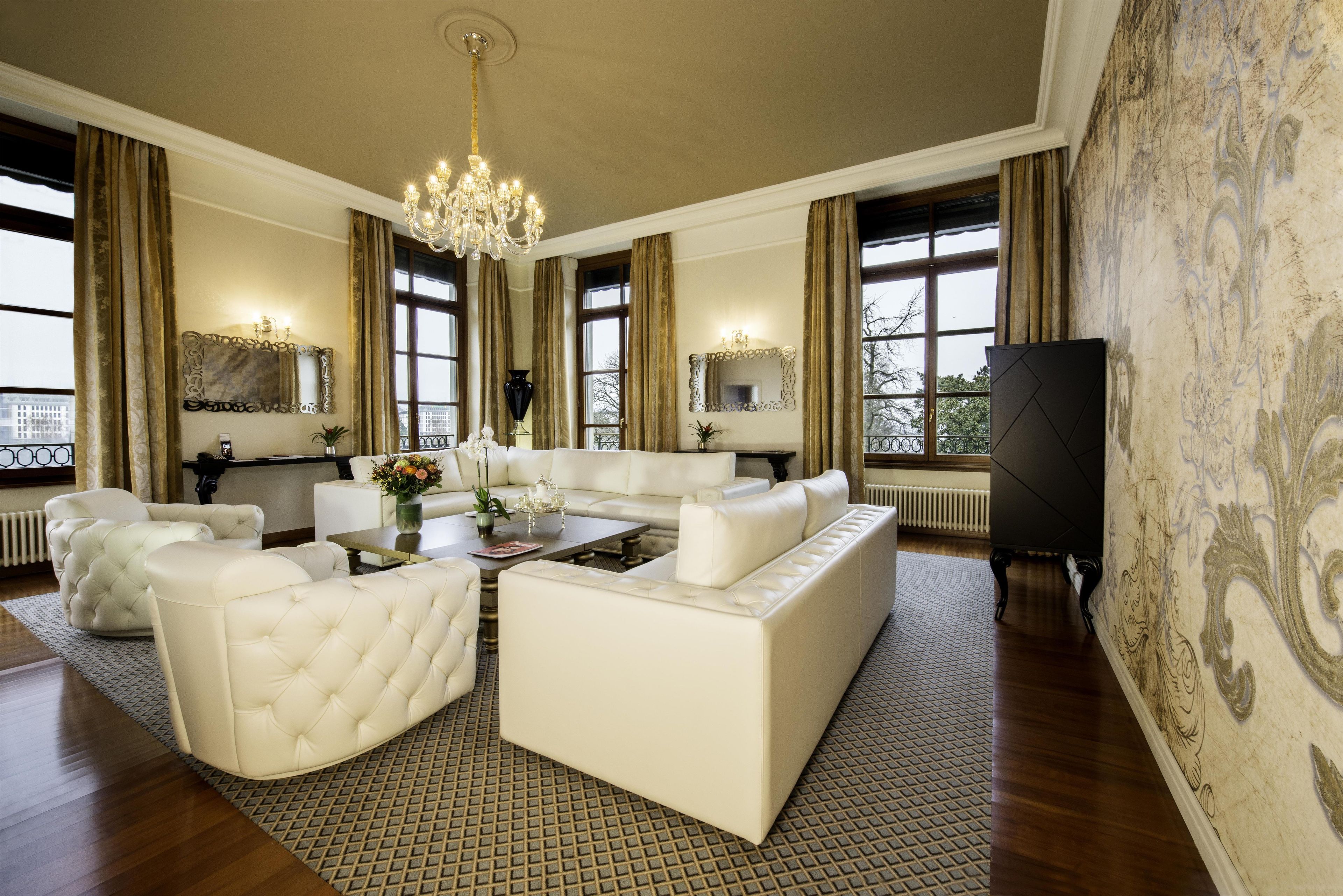 A spacious living room with a large white sofa, a coffee table, and a chandelier. There are two large windows with curtains, a fireplace, and a television on the right.