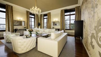 A spacious living room with a large white sofa, a coffee table, and a chandelier. There are two large windows with curtains, a fireplace, and a television on the right.