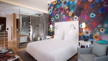 Une chambre d’hôtel moderne dotée d’un grand lit, d’une douche à parois de verre, d’une fresque murale aux motifs floraux éclatants et d’une petite table ronde avec une lampe.