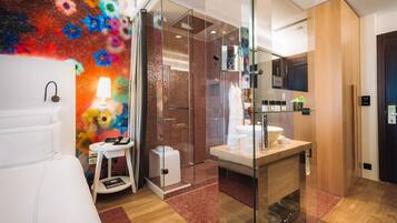 A modern hotel room with a bed, a glass shower enclosure, a wooden vanity, and a colorful abstract wall.