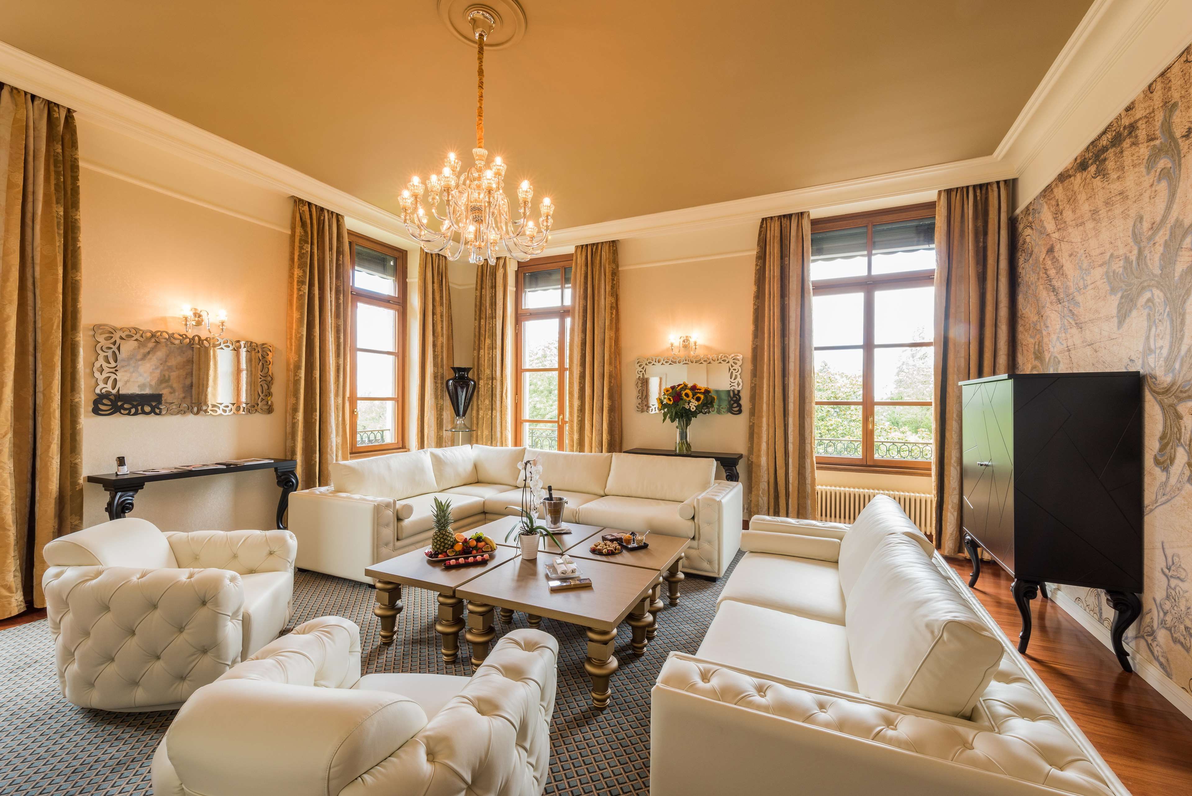 A spacious living room with a large chandelier, white leather sofas, a coffee table with fruit, and a television on the right.