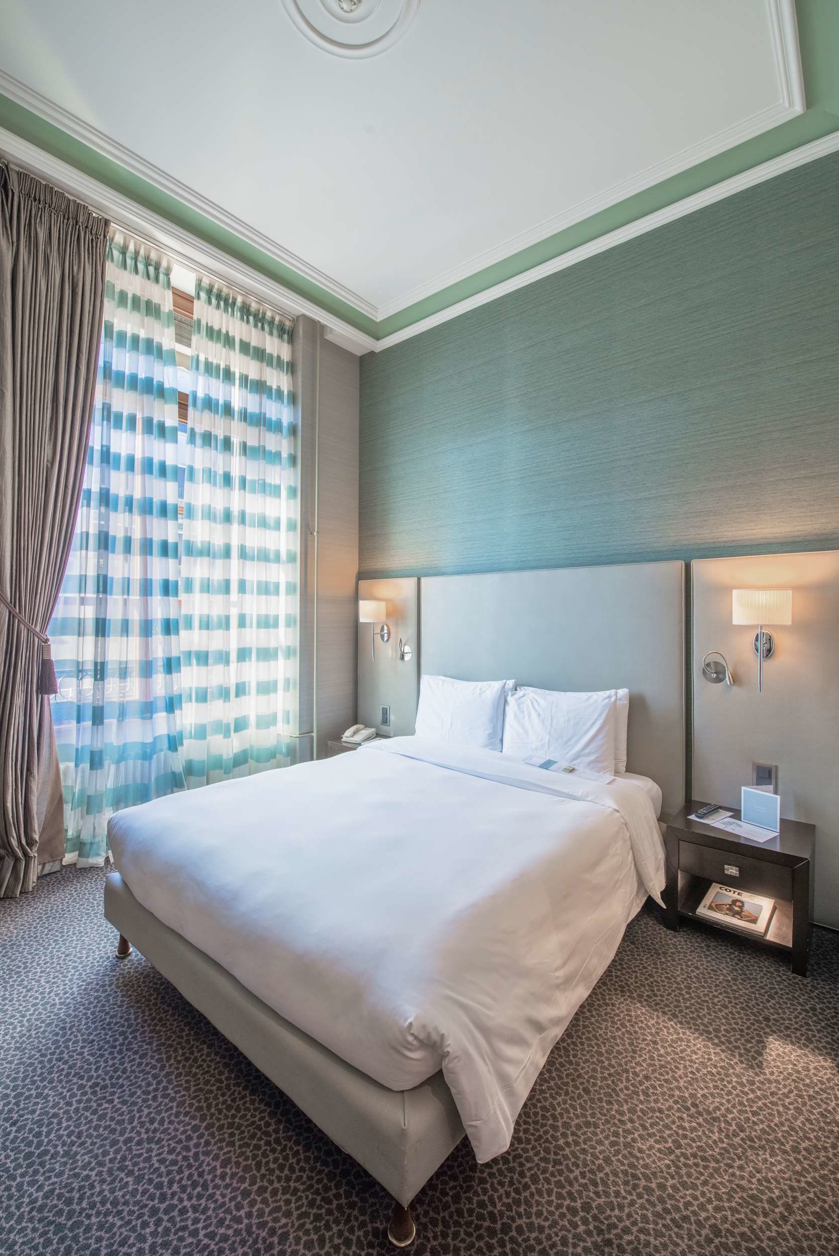 Una habitación de hotel con una cama grande, una mesita de noche, una lámpara y una ventana con cortinas.