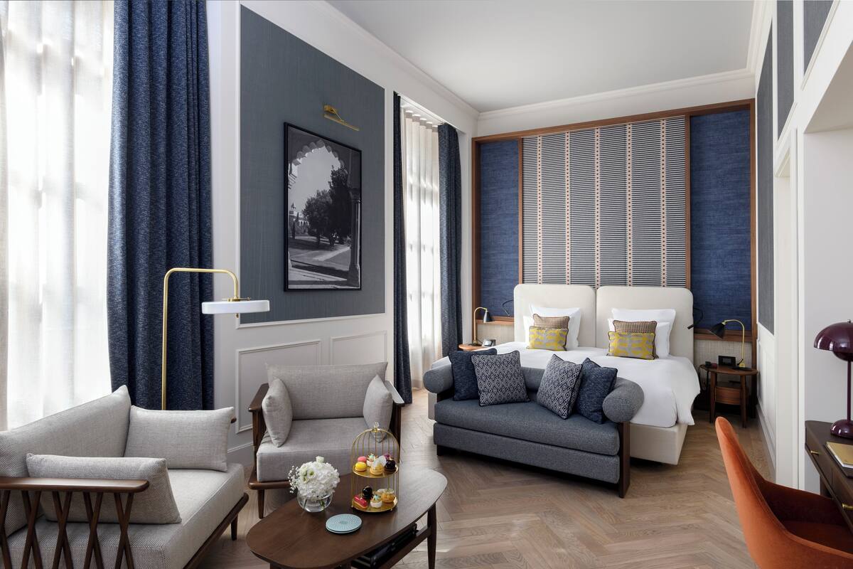 Una habitación de hotel moderna con un sofá gris, un sillón más pequeño, una mesa de centro de madera con un florero y una bandeja de pasteles, una cama con dos almohadas, una mesita de noche con una lámpara y un ventanal con persianas.