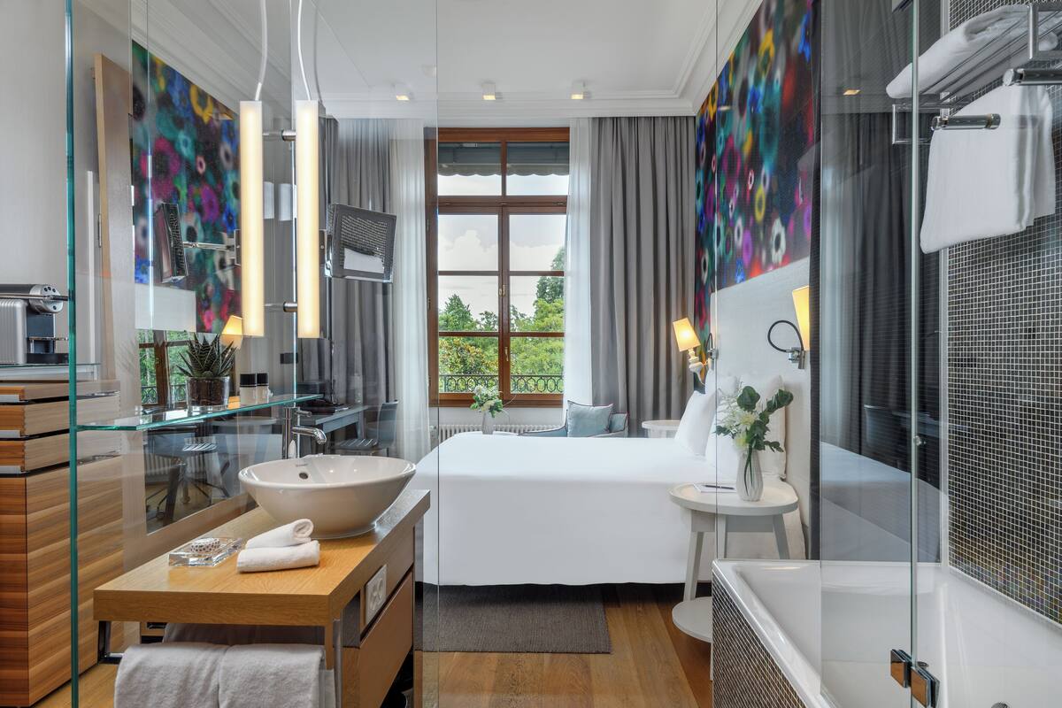 Habitación de hotel moderna con una cama grande, un baño con paredes de cristal, una cómoda de madera y vistas a zonas verdes.