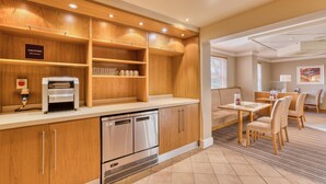 Una cocina de hotel con armarios de madera, una cafetera, un lavavajillas y un comedor con una mesa y sillas.