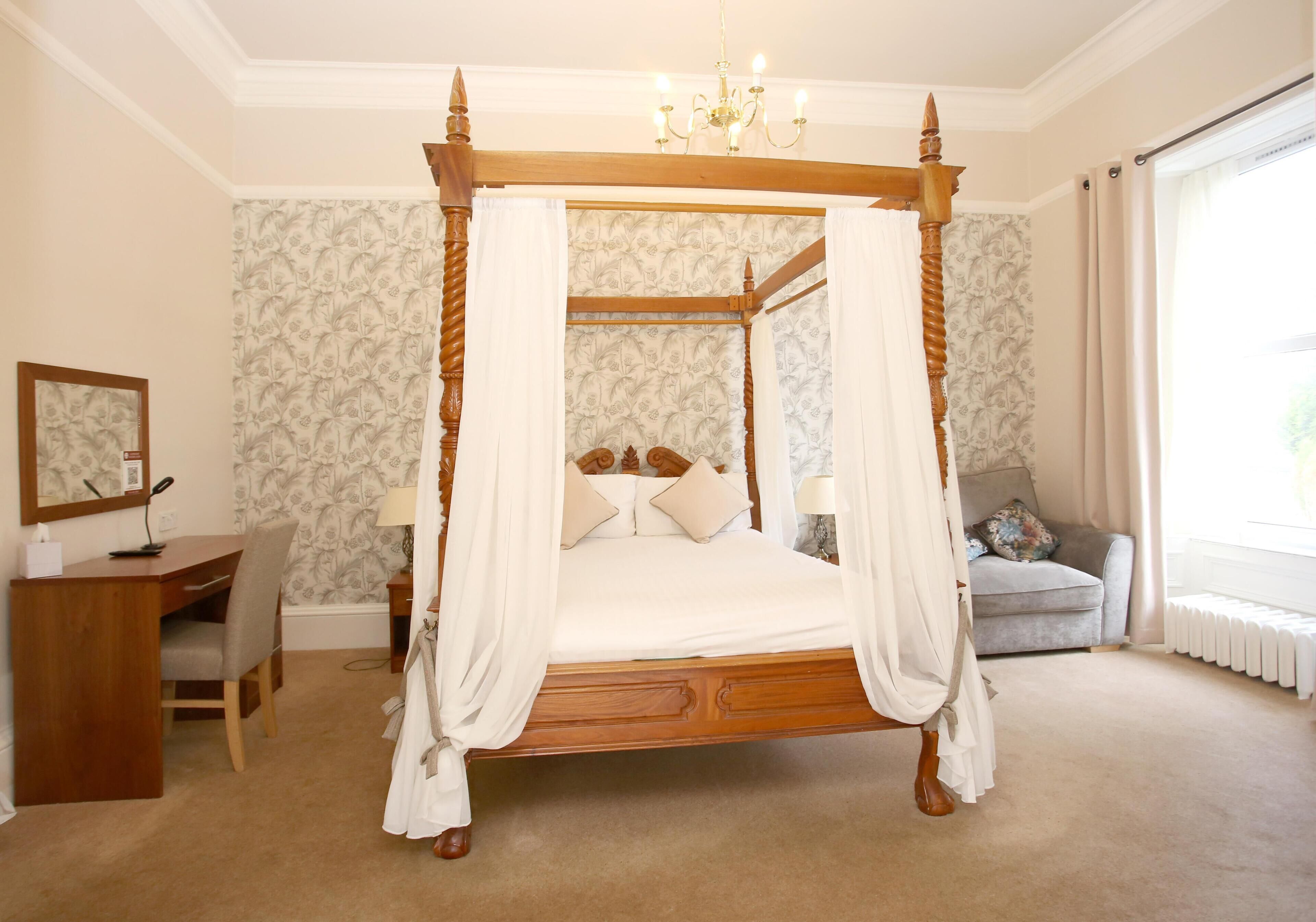 Een bed met hemelbed, met houten frame en witte beddengoed, een bureau met stoel, een kleine tafel met lamp, en een bank met kussens.