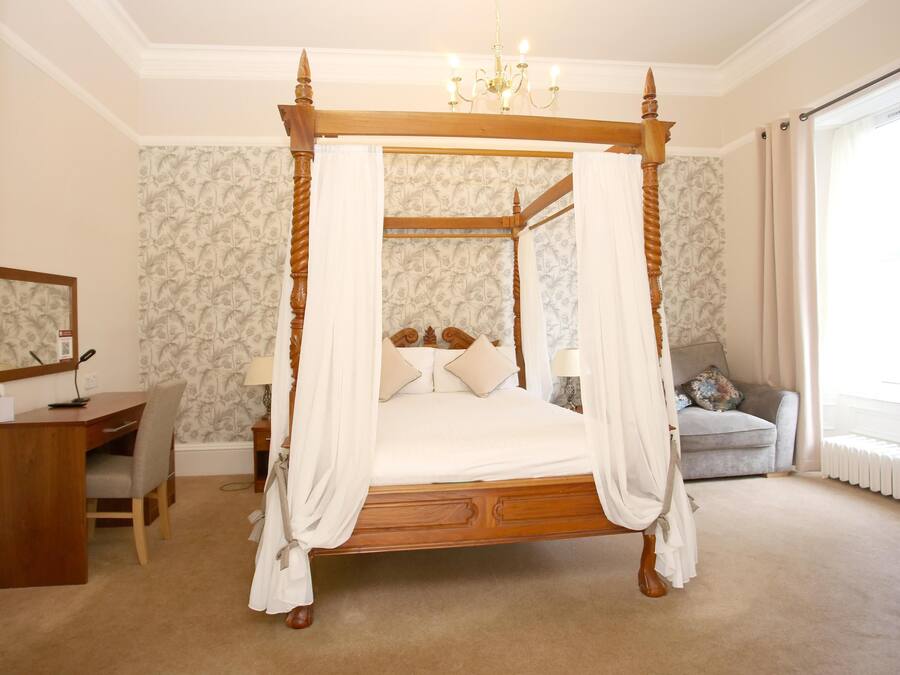 A four-poster canopy bed with a wooden frame and white bedding, a desk with a chair, a small table with a lamp, and a sofa with cushions.
