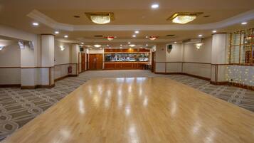 A spacious ballroom with a polished wooden floor, a bar area with a variety of bottles, and a chandelier.
