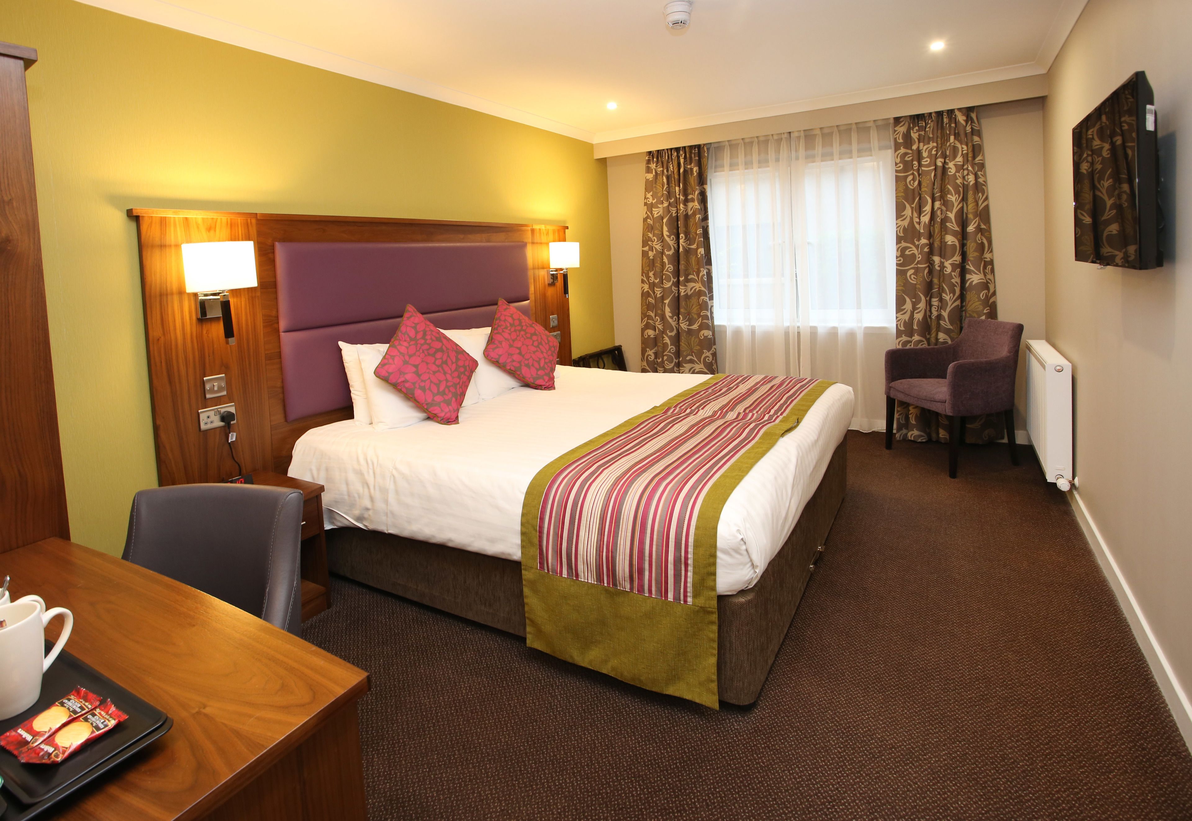 A hotel room with a large bed, a desk with a chair, a TV, and a window with curtains.