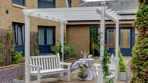 Une terrasse couverte avec des bancs blancs, une petite table agrémentée d’un arrangement floral et une plante en pot.