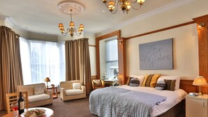 Une chambre d’hôtel avec un grand lit, un bureau, une chaise, une table à manger avec une assiette de nourriture et une bouteille de vin, et un lustre.