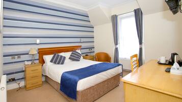 Une chambre d’hôtel avec un lit, un bureau en bois, une chaise, une lampe et une fenêtre avec des rideaux.