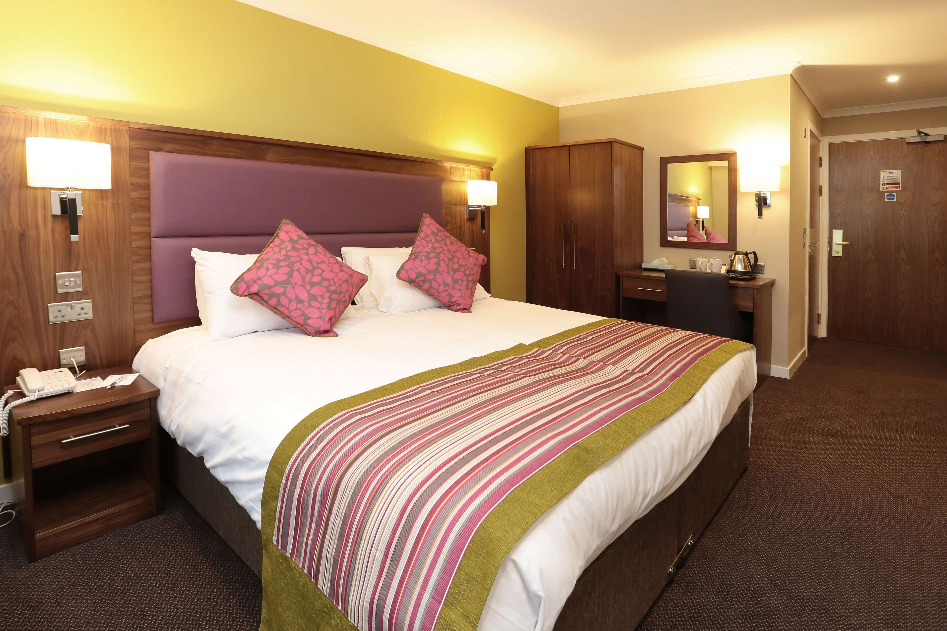 A hotel room with a large bed, wooden headboard, two bedside lamps, a nightstand with a phone, a desk with a mirror, and a wardrobe.