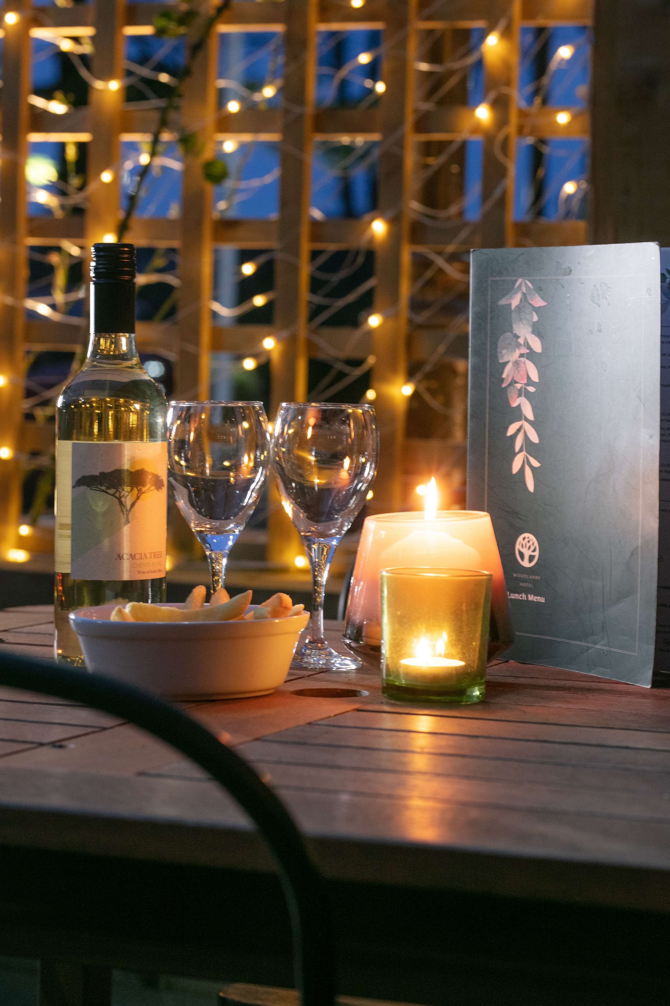 A table with a bottle of wine, two wine glasses, a lit candle, a menu, and a bowl of snacks.