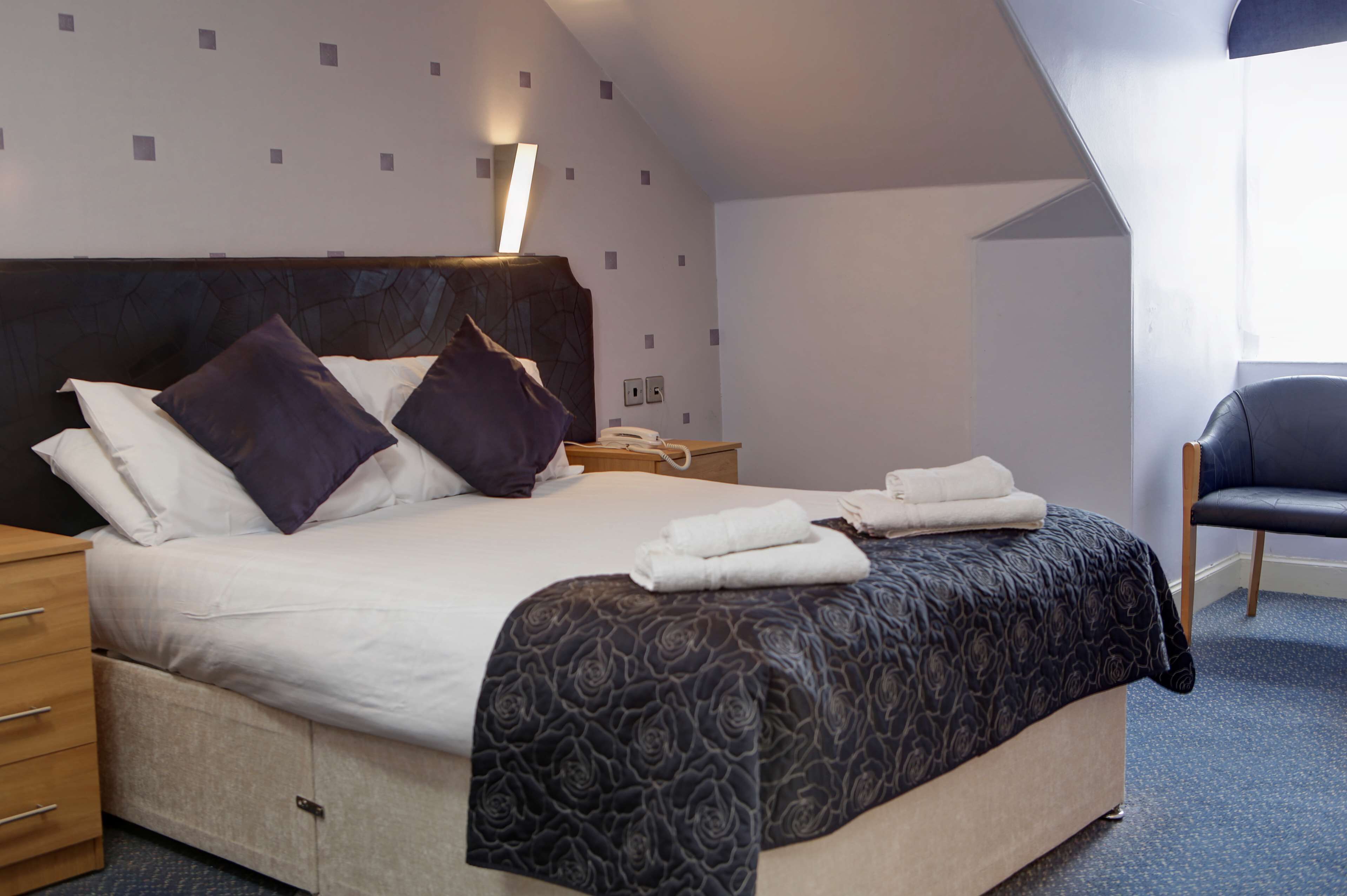A neatly made bed with a dark patterned bedspread, a bedside table with a lamp, and a chair in a corner.