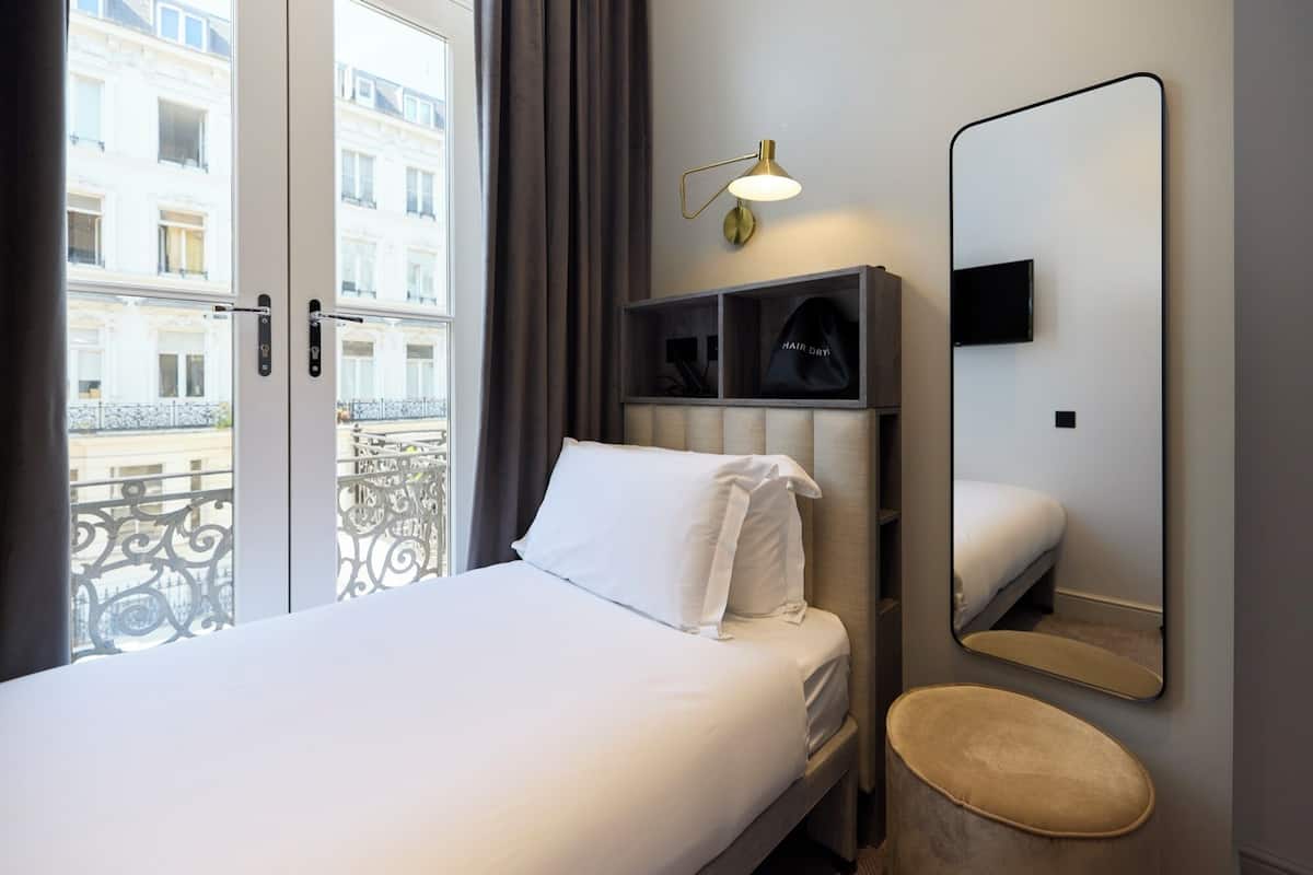 A hotel room with a bed, a bedside table, a mirror, a wall-mounted TV, and a view of a balcony with a railing.