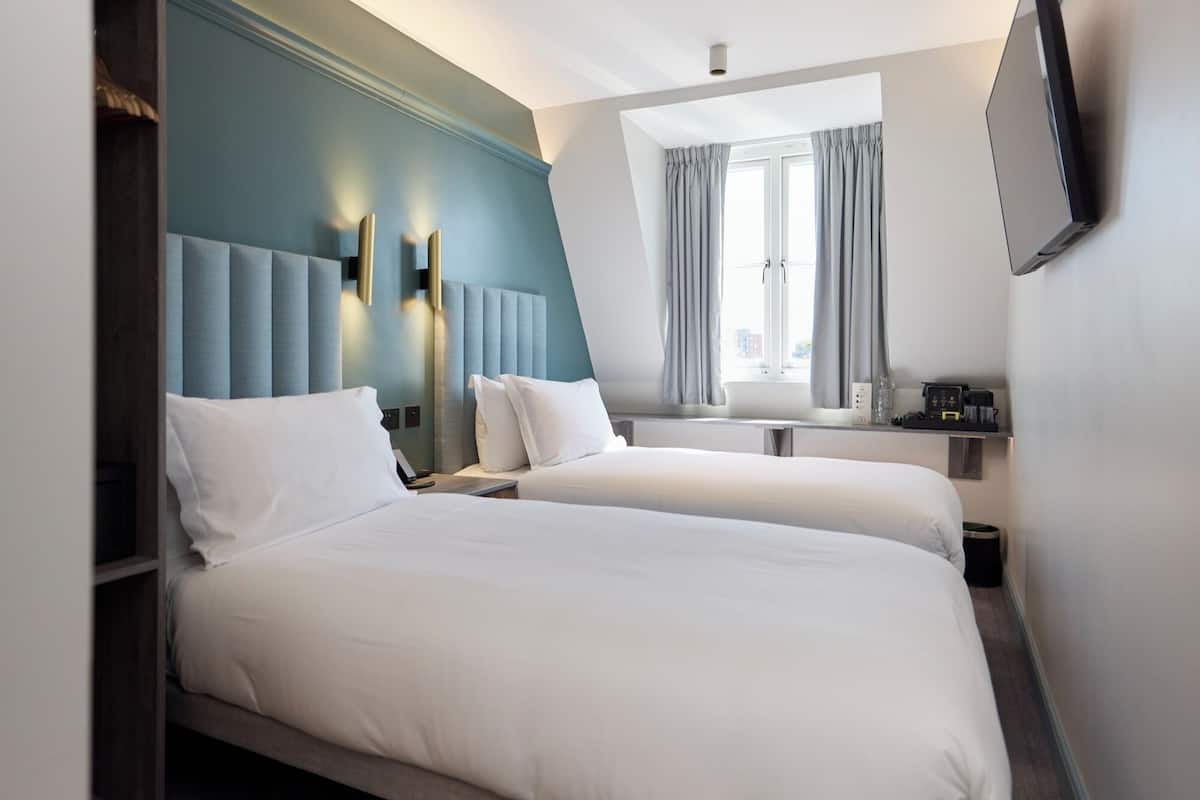 A hotel room with two beds, a large window with curtains, a flat-screen TV mounted on the wall, and a small table with a coffee machine.