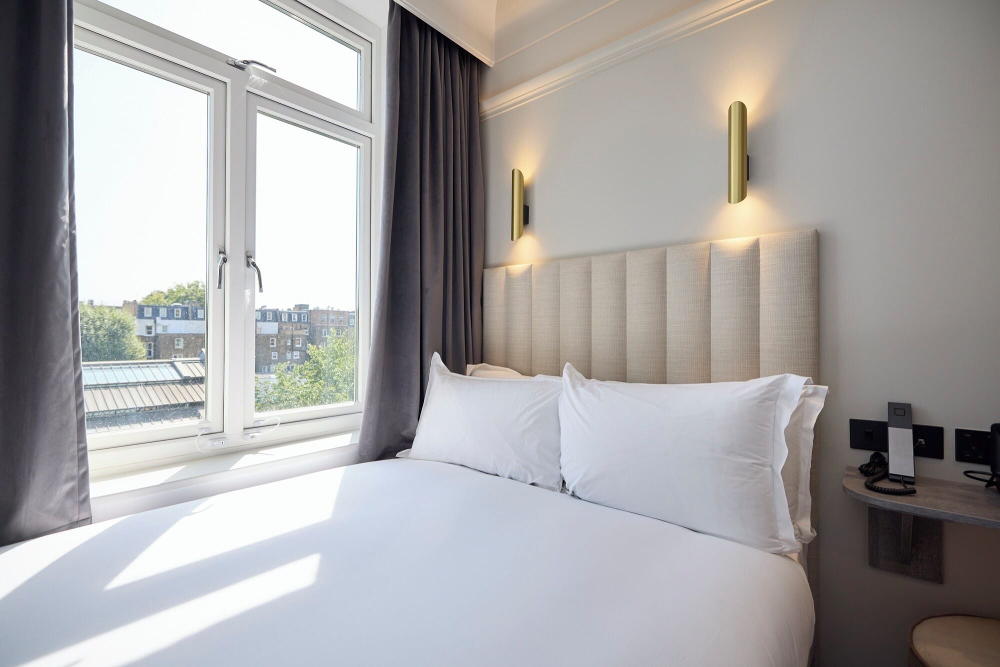 A hotel room with a large bed, a window with curtains, a headboard with wall-mounted lights, and a small bedside table with a phone and a lamp.